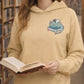 Sand coloured hoodie with a dragon laying on a book on the left chest, worn by a female reading a book in a library. 