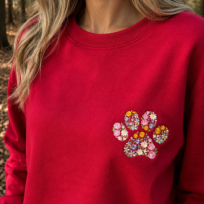 A close-up of a woman wearing a bright red crewneck sweatshirt with a colorful embroidered floral paw print on the left chest, set against a blurred outdoor forest background.