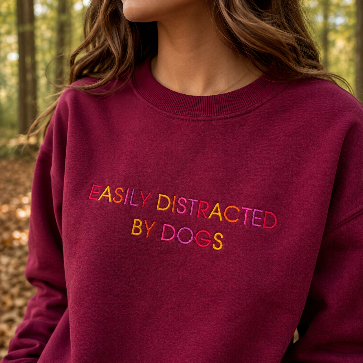 A woman wearing a burgundy crewneck sweatshirt with the phrase 'EASILY DISTRACTED BY DOGS' embroidered in multicolored letters, set against a blurred forest background.