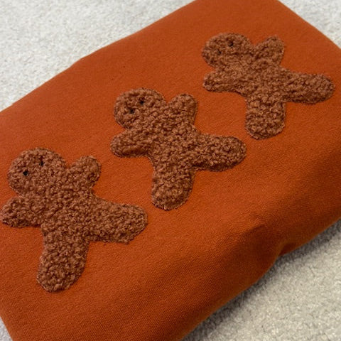 Three brown gingerbread men appliqued onto a ginger coloured sweatshirt.
