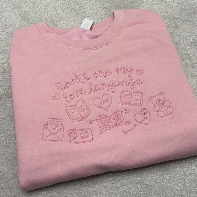 Pink sweatshirt with embroidered text and graphics saying Books are my love language. Images of love letters and teddy bear