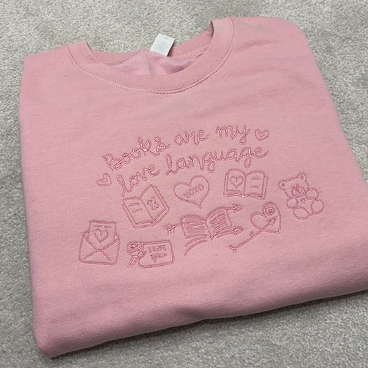 Pink sweatshirt with embroidered text and graphics saying Books are my love language. Images of love letters and teddy bear