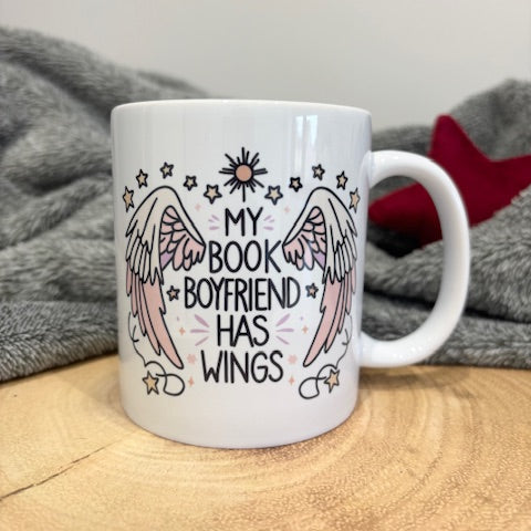 My Book Boyfriend Has Wings printed on white ceramic mug, shown on a wooden worktop with a grey blanket behind.
