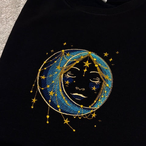 Black sweatshirt with a celestial design featuring a face within a moon and stars.