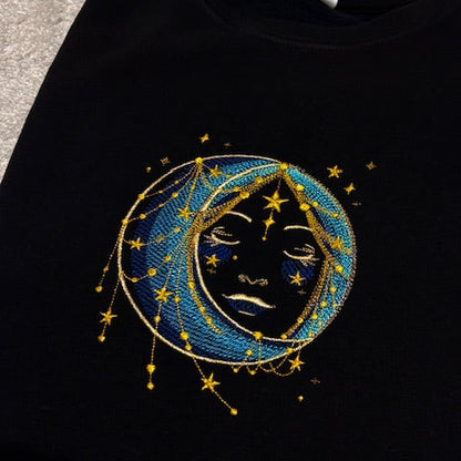 Black sweatshirt with a celestial design featuring a face within a moon and stars.