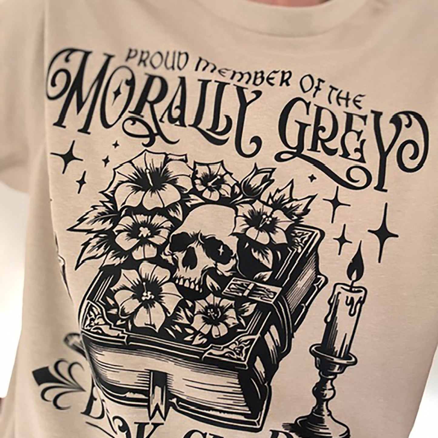 Beige sweatshirt with black graphic design featuring a skull, book, and candle, text 'Proud Member of the Morally Grey Dark Club Weekly'.