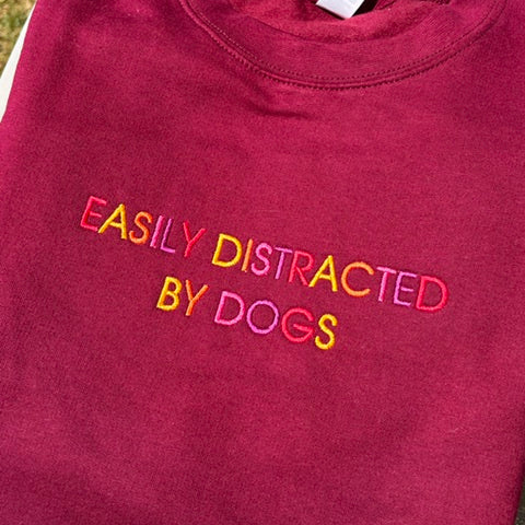 A folded maroon crewneck sweatshirt with the phrase 'EASILY DISTRACTED BY DOGS' embroidered across the chest in a multi-colored gradient of pink, yellow, orange, and green.
