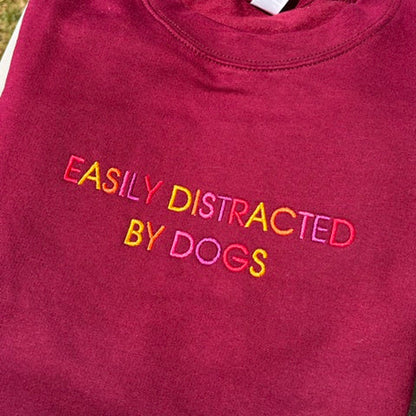 A folded maroon crewneck sweatshirt with the phrase 'EASILY DISTRACTED BY DOGS' embroidered across the chest in a multi-colored gradient of pink, yellow, orange, and green.
