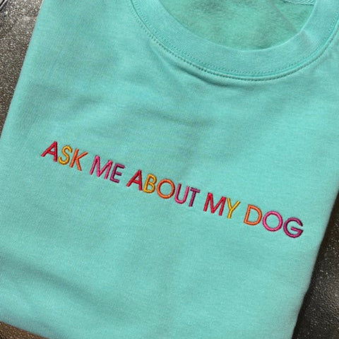A folded mint green t-shirt with the phrase 'ASK ME ABOUT MY DOG' embroidered across the chest in a vibrant rainbow gradient.