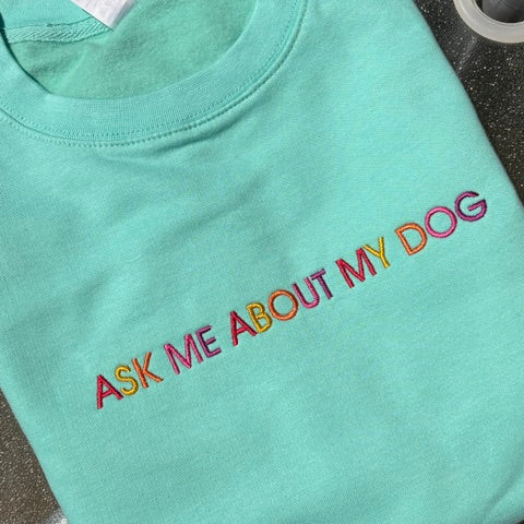 A mint green t-shirt with the phrase 'ASK ME ABOUT MY DOG' embroidered in a rainbow gradient across the chest, laid flat on a dark textured surface.