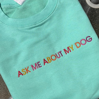 A mint green t-shirt with the phrase 'ASK ME ABOUT MY DOG' embroidered in a rainbow gradient across the chest, laid flat on a dark textured surface.