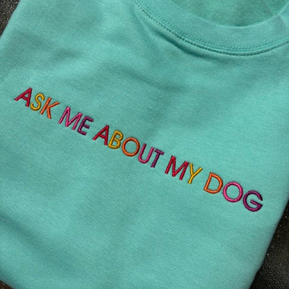 A mint green crewneck t-shirt with the phrase 'ASK ME ABOUT MY DOG' embroidered across the chest in a rainbow gradient. The shirt is laid flat on a dark textured surface, showing a white brand tag at the collar.