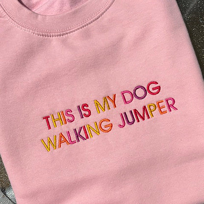 A folded light pink crewneck sweatshirt featuring the text 'THIS IS MY DOG WALKING JUMPER' embroidered in colorful pink, orange, yellow, and red letters.