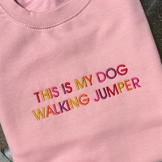 A folded light pink crewneck sweatshirt featuring the text 'THIS IS MY DOG WALKING JUMPER' embroidered in colorful pink, orange, yellow, and red letters.