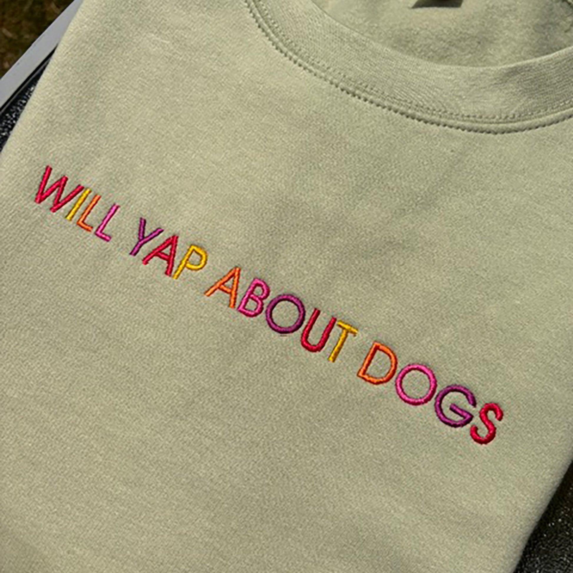 A close-up of a light sage green t-shirt featuring the phrase 'WILL YAP ABOUT DOGS' embroidered across the chest in a vibrant multicolor gradient.