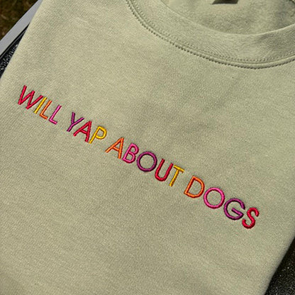 A close-up of a light sage green t-shirt featuring the phrase 'WILL YAP ABOUT DOGS' embroidered across the chest in a vibrant multicolor gradient.