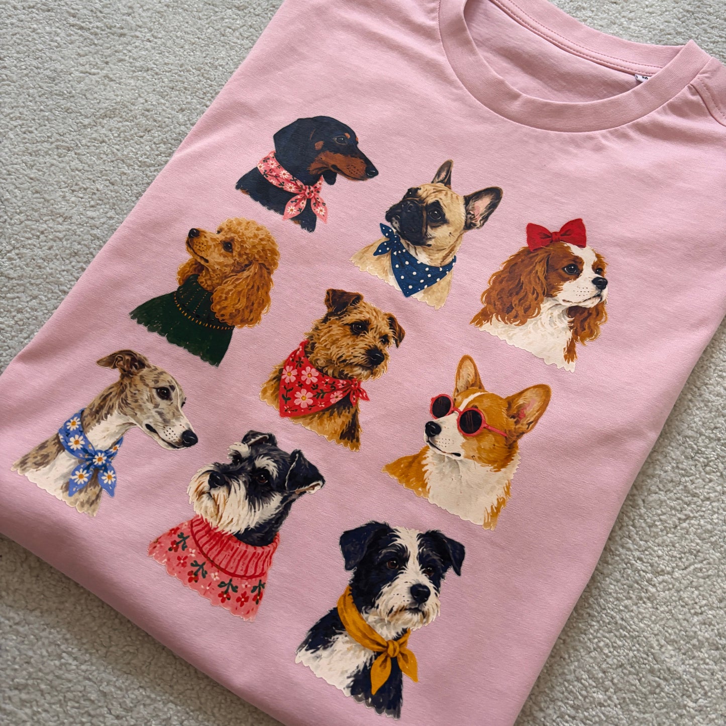 A light pink short-sleeve t-shirt laid flat on a textured gray surface, featuring a graphic print of nine different dog breeds wearing colorful bandanas and sweaters.