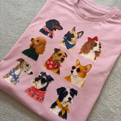 A light pink short-sleeve t-shirt laid flat on a textured gray surface, featuring a graphic print of nine different dog breeds wearing colorful bandanas and sweaters.