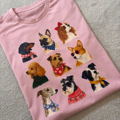 A folded light pink crewneck t-shirt featuring a graphic print of nine different dog illustrations wearing colorful sweaters and accessories, laid flat on a textured gray carpet.