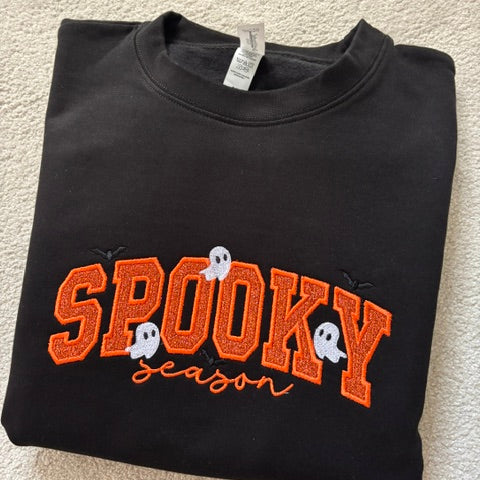 A folded black crewneck sweatshirt featuring a 'SPOOKY season' graphic in orange glitter-style lettering with white ghosts and small black spider details, laid flat on a beige carpet.