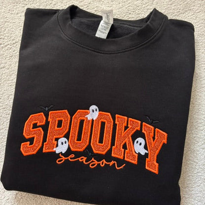 A folded black crewneck sweatshirt featuring a 'SPOOKY season' graphic in orange glitter-style lettering with white ghosts and small black spider details, laid flat on a beige carpet.