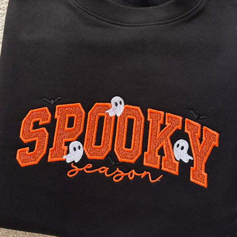 A folded black crewneck sweatshirt featuring 'SPOOKY season' embroidered in orange and white on the front, resting on a beige carpet.