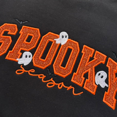 Close-up of a black garment with orange embroidered 'SPOOKY' lettering, small white ghosts, and the word 'season' in orange script.
