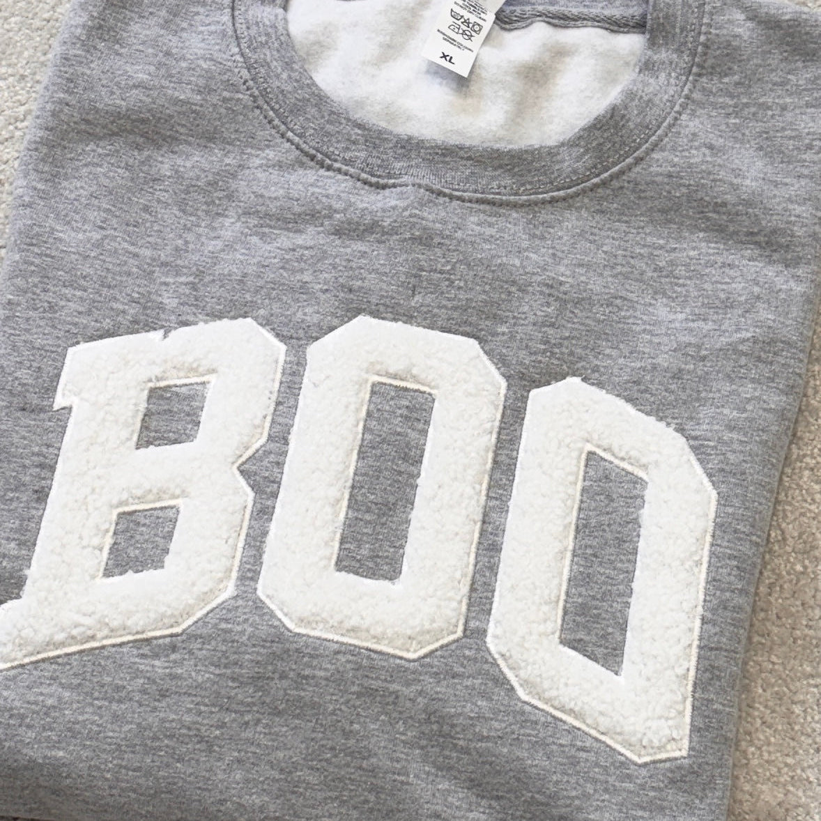 A folded heather gray sweatshirt with large white varsity-style 'BOO' lettering across the front, placed on a beige carpeted floor.