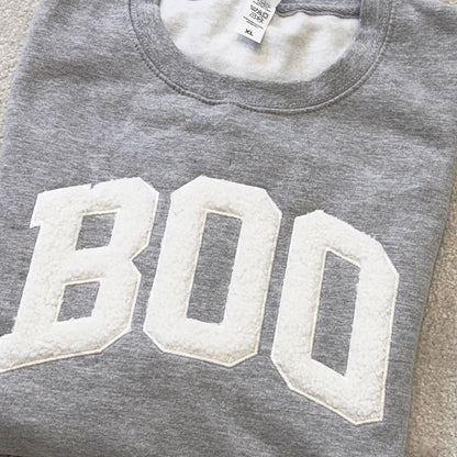 A folded heather gray sweatshirt with large white varsity-style 'BOO' lettering across the front, placed on a beige carpeted floor.