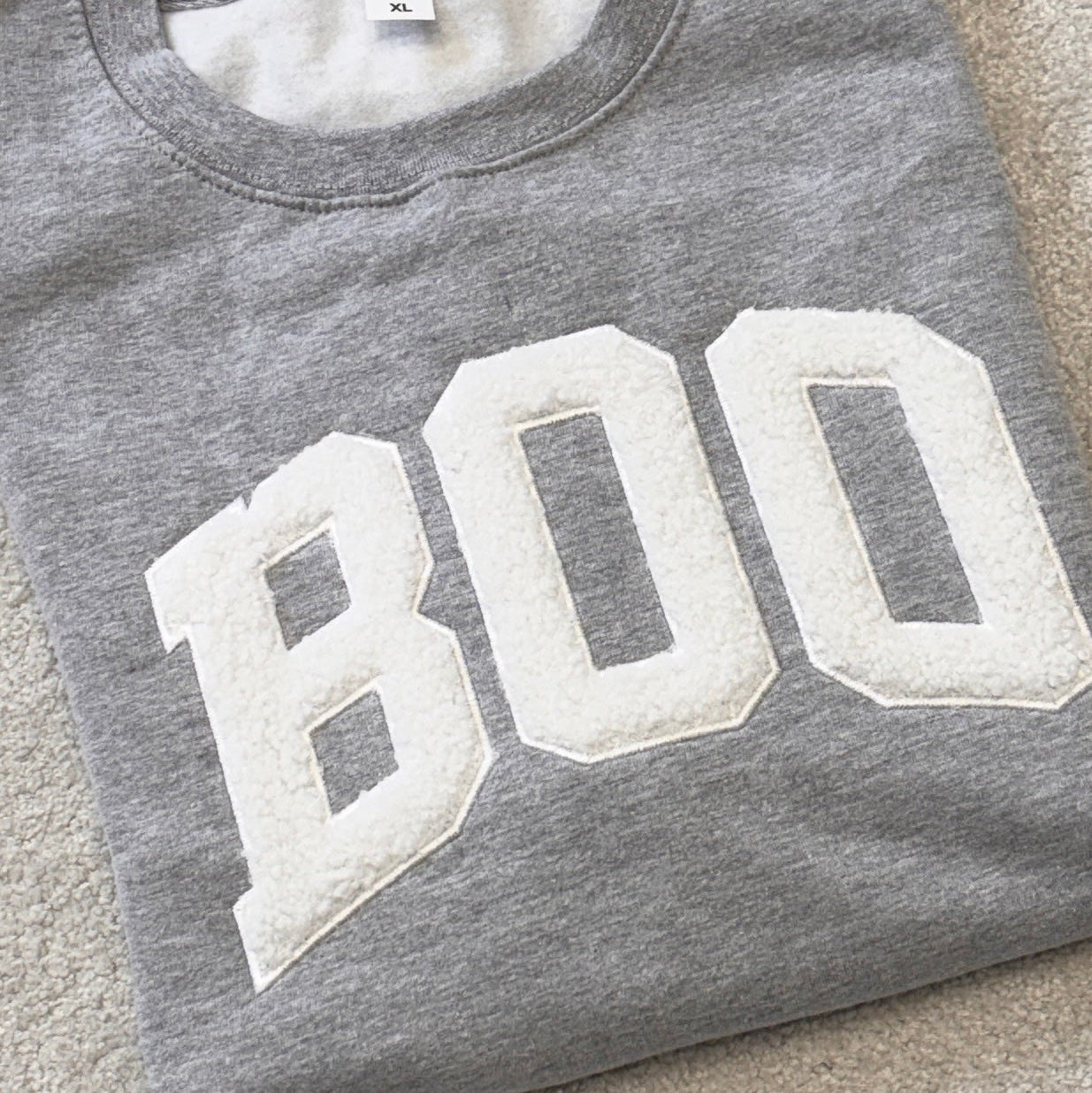A folded heather gray sweatshirt with large white varsity-style letters spelling 'BOO' across the front, laid flat on a light beige carpet.