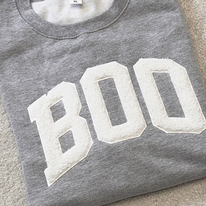 A folded heather gray sweatshirt with large white varsity-style letters spelling 'BOO' across the front, laid flat on a light beige carpet.