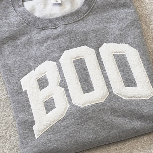 A folded heather gray sweatshirt with large white varsity-style letters spelling 'BOO' across the front, laid flat on a light beige carpet.