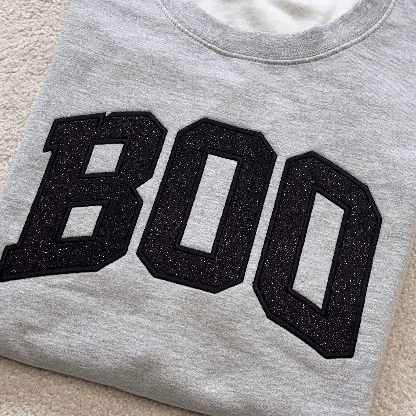 A folded light gray sweatshirt featuring the word 'BOO' in large black glittery varsity-style letters across the front, laid flat on a beige textured carpet.