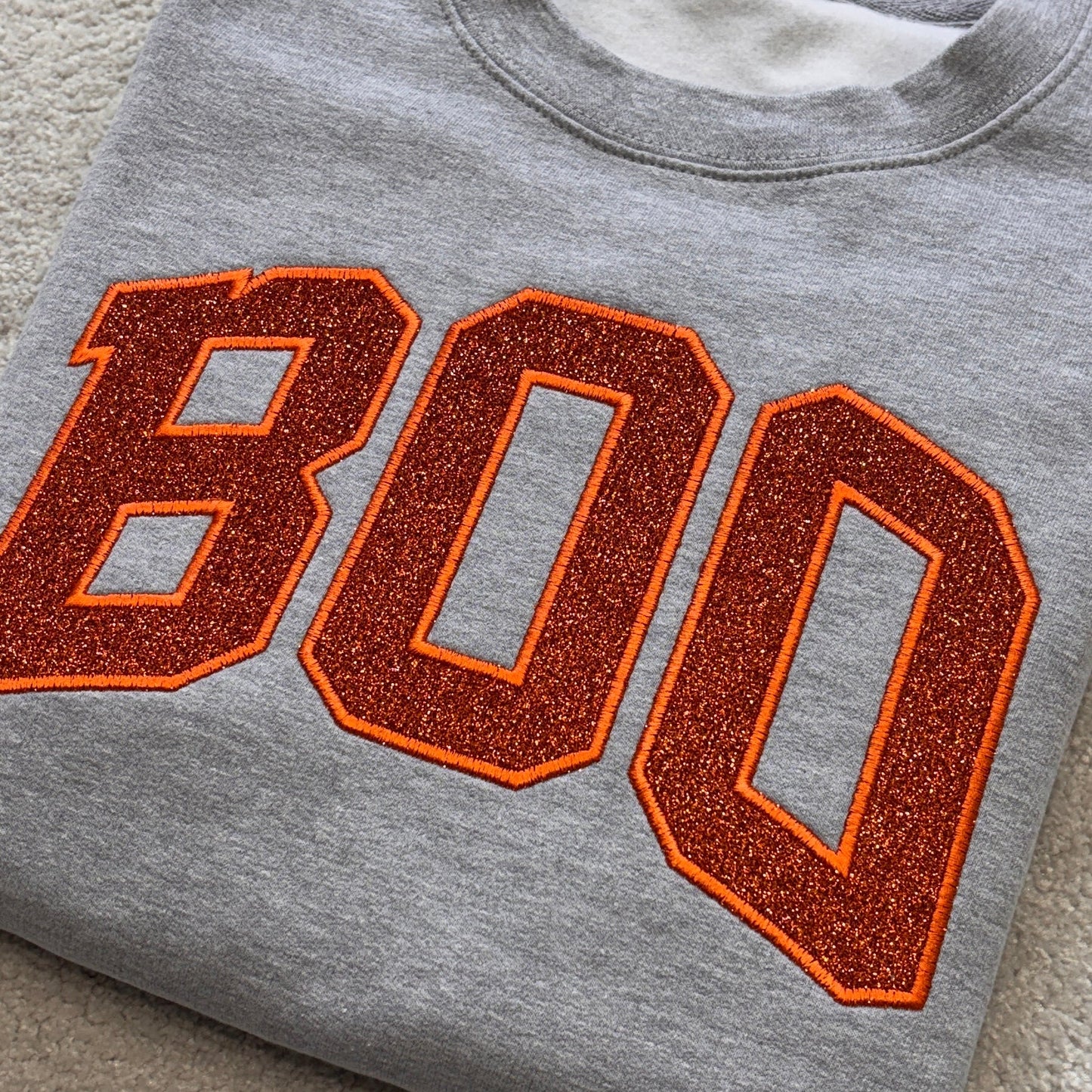 A folded heather gray crewneck sweatshirt featuring the word 'BOO' in large orange varsity-style letters with a glittery orange-brown fill and a thin orange outline, laid flat on a beige carpet.