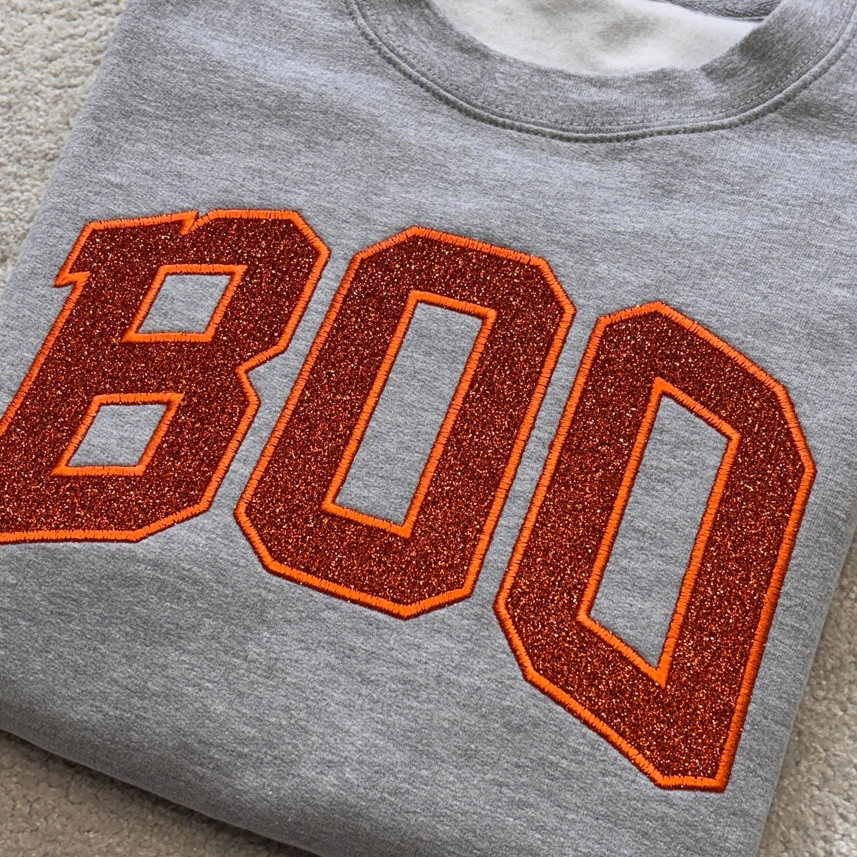A folded heather gray crewneck sweatshirt featuring the word 'BOO' in large orange varsity-style letters with a glittery orange-brown fill and a thin orange outline, laid flat on a beige carpet.