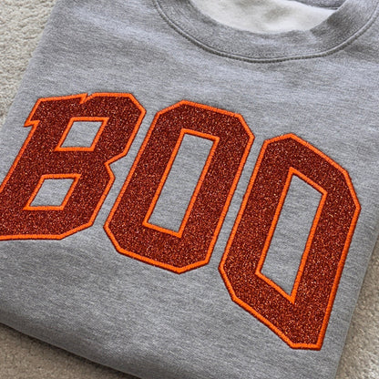 A folded heather gray crewneck sweatshirt featuring the word 'BOO' in large orange varsity-style letters with a glittery orange-brown fill and a thin orange outline, laid flat on a beige carpet.