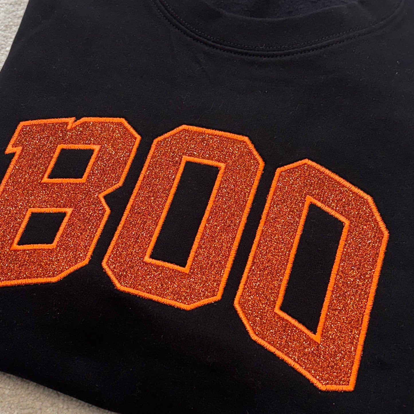 A folded black crewneck sweatshirt featuring the word 'BOO' in large orange varsity-style letters with a glittery texture and orange outline, laid flat on a light beige textured surface.