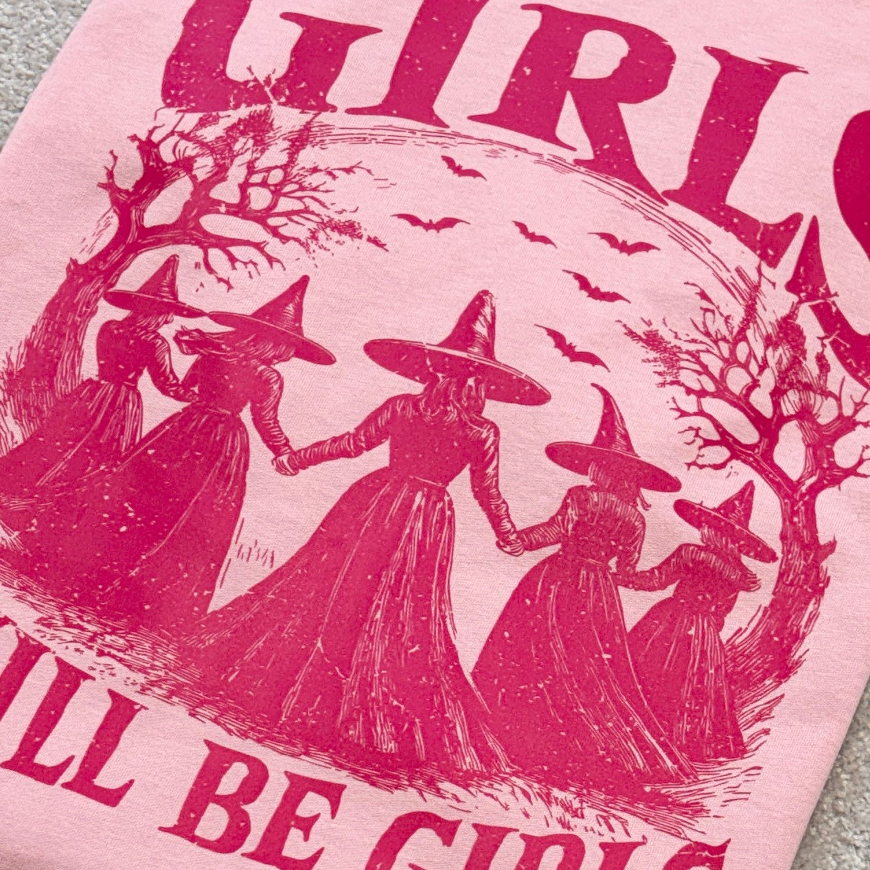 A close-up of a light pink t-shirt featuring a dark pink graphic print of four witches holding hands inside a circle, with the text 'GIRLS WILL BE GIRLS' printed around them.