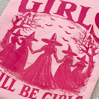 A close-up of a light pink t-shirt featuring a dark pink graphic print of four witches holding hands inside a circle, with the text 'GIRLS WILL BE GIRLS' printed around them.