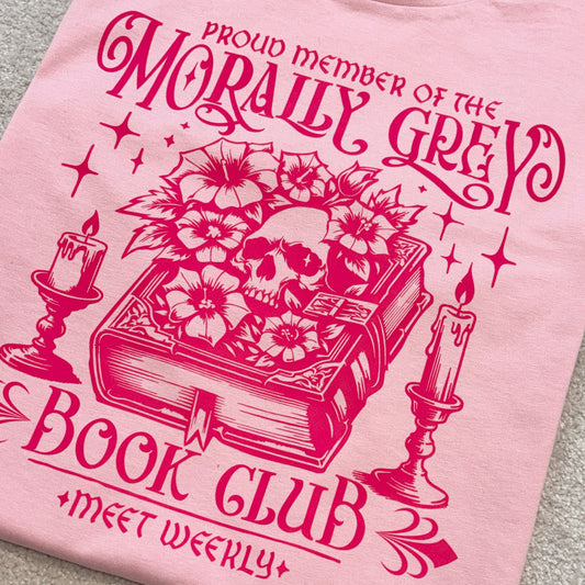 A light pink crewneck t-shirt laid flat on a gray carpet, featuring a dark pink graphic print of a skull in a book surrounded by flowers, candles, and gothic text that reads 'PROUD MEMBER OF THE MORALLY GREY BOOK CLUB MEET WEEKLY'.