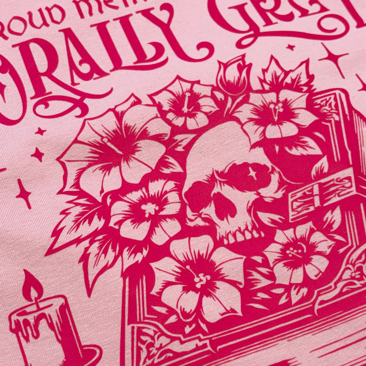 A close-up of a pink graphic t-shirt featuring a dark red screen-printed design of a skull surrounded by hibiscus flowers, with the text 'PROUD MEMBER OF THE ORALLY GREVED' arched above.