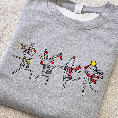 A folded heather gray crewneck sweatshirt featuring a graphic of four dancing reindeer decorated with holiday lights, red nose accents, and scarves, laid flat on a beige carpet.