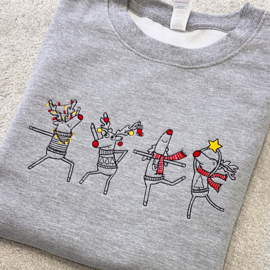 A folded heather gray crewneck sweatshirt featuring a graphic of four dancing reindeer decorated with holiday lights, red nose accents, and scarves, laid flat on a beige carpet.