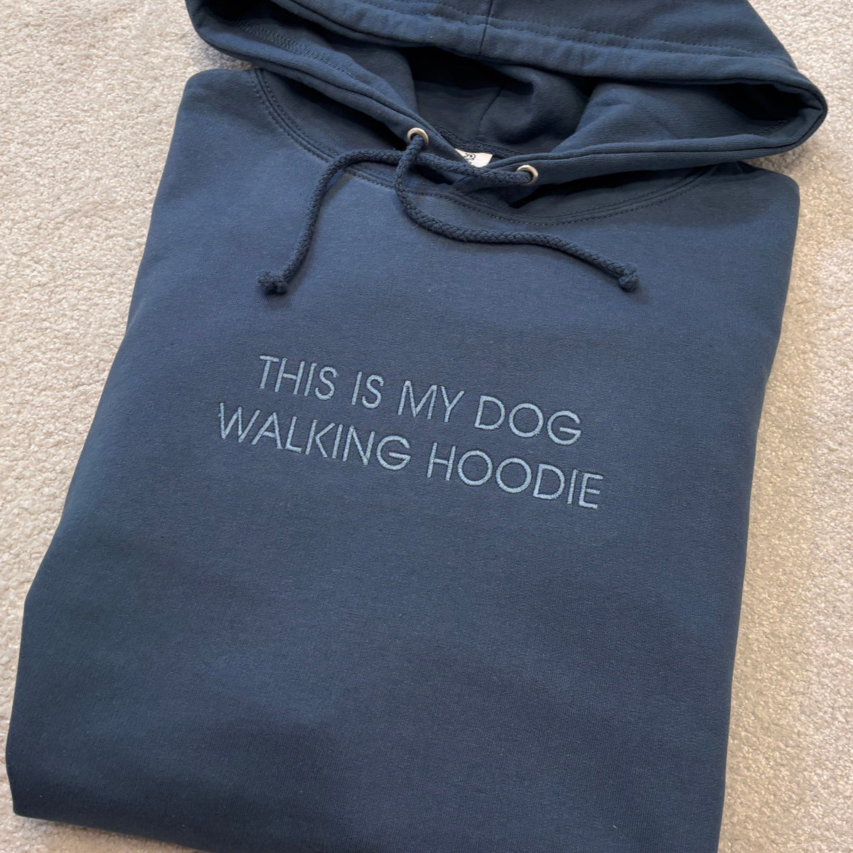 THIS IS MY DOG WALKING HOODIE embroidered in blue thread on an airforce blue hoodie which is folded and laying on a carpet