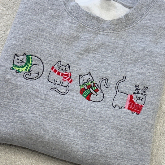 Four sketch style cats across the front of a heather grey sweatshirt, wearing christmas jumpers and scarves