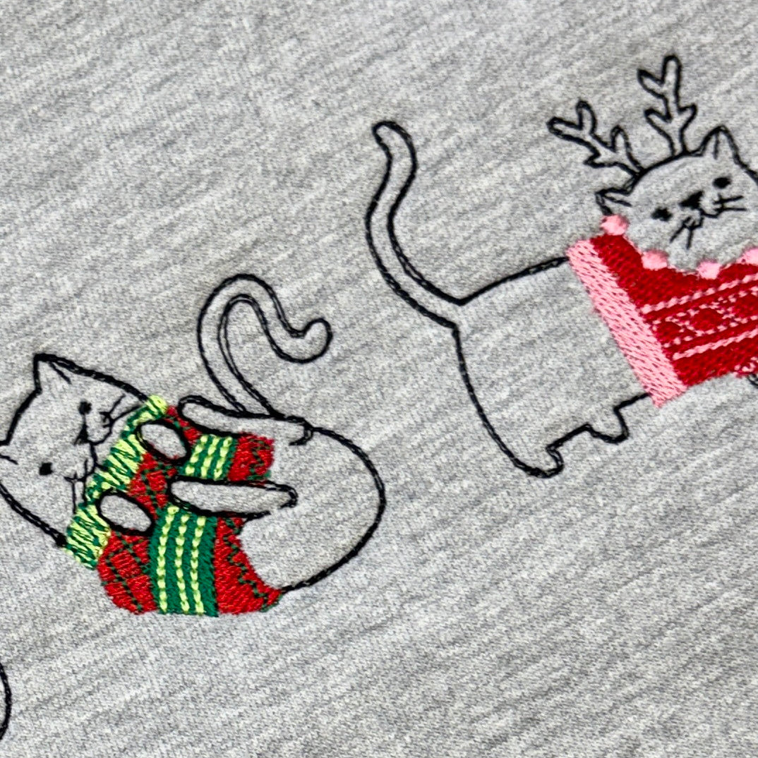 Close-up of a heather gray fabric with embroidered cartoon cats wearing colorful knit sweaters, including green and red stripes and a red sweater with reindeer antlers.