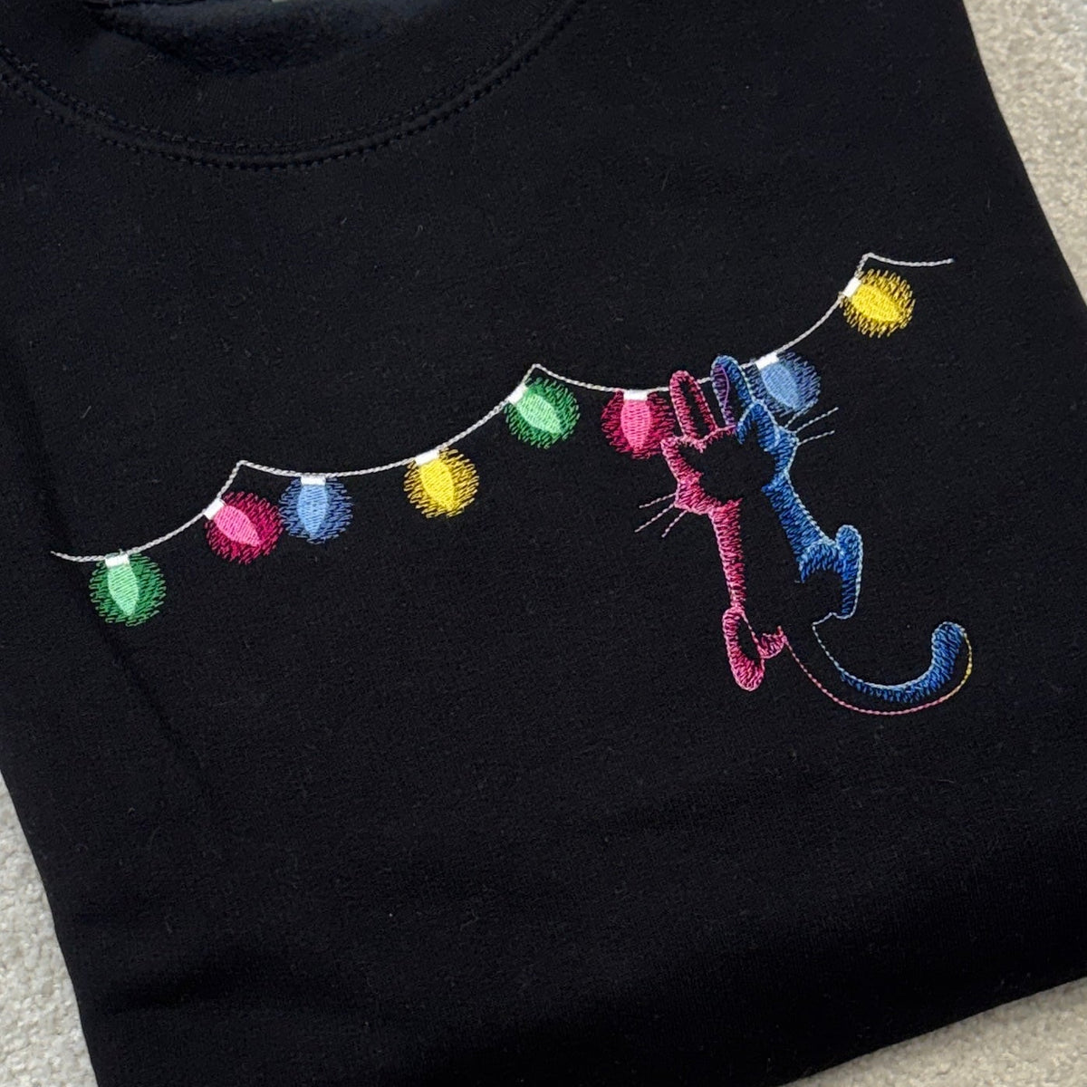 A folded black crewneck sweatshirt featuring a colorful embroidered design of two cartoon cats hanging from a string of multicolored Christmas lights, laid flat on a light beige textured surface.