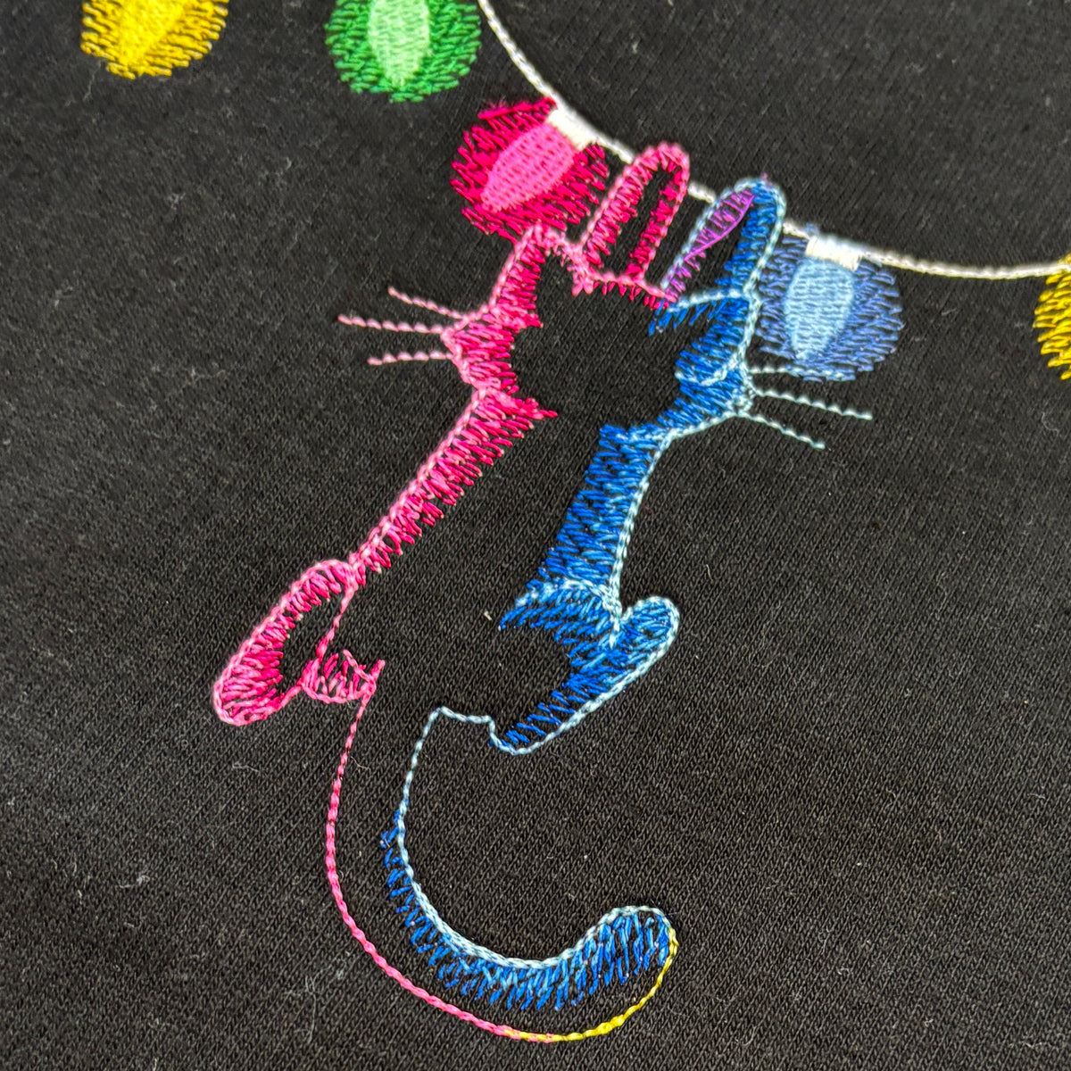 Close-up of embroidered black cats reaching up toward colorful string lights on a black fabric background.