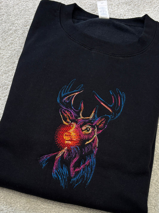 A folded black crewneck sweatshirt featuring a colorful embroidered reindeer graphic on the front, laid flat on a light gray textured surface.
