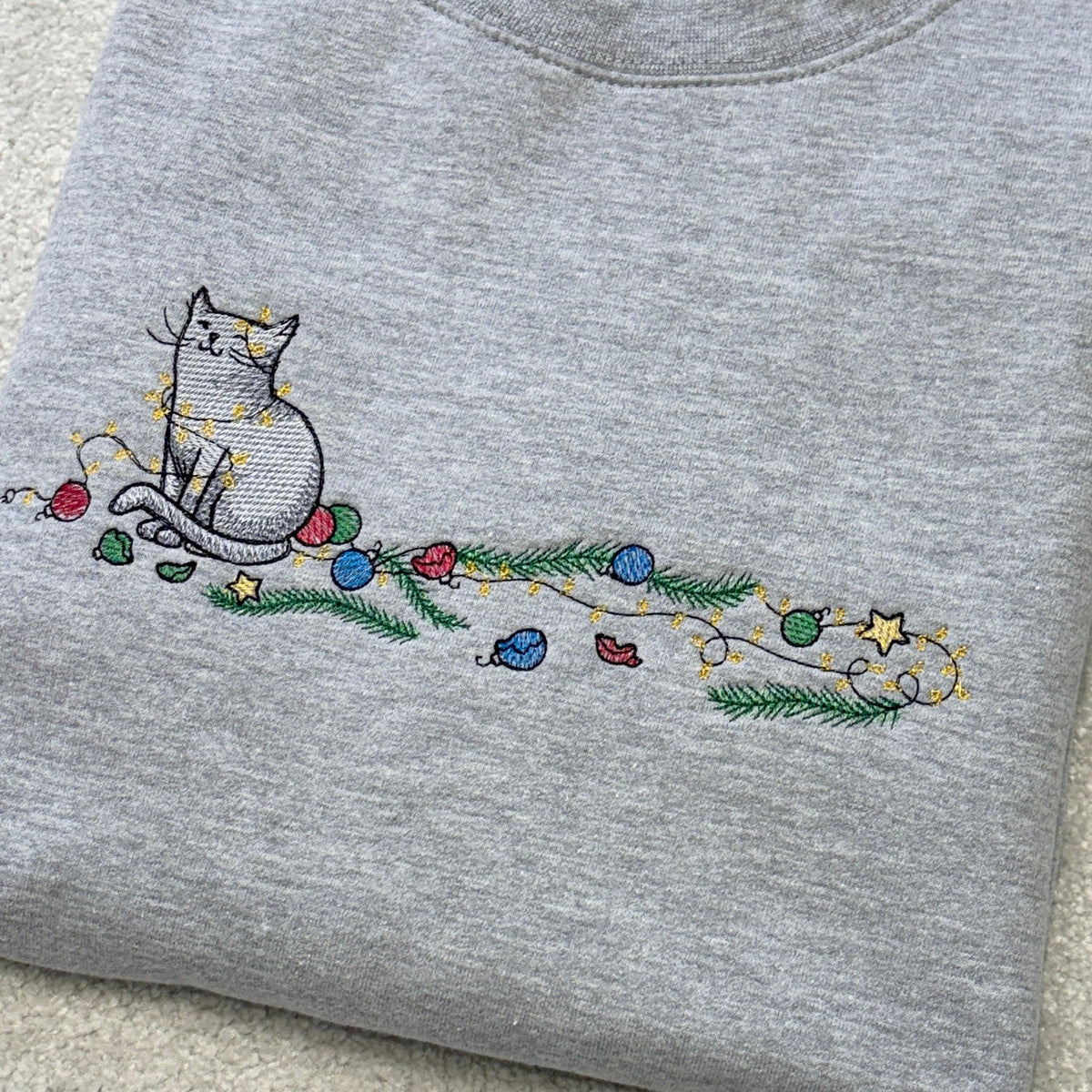 A folded heather gray crewneck sweatshirt featuring a colorful embroidered design of a cat wrapped in holiday lights and pine branches, laid flat on a light beige textured surface.